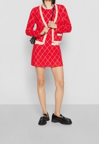 Mujer vestida con una falda roja de patrón de diamantes y un cárdigan a juego con ribete crema, sosteniendo un bolso cruzado negro y zapatos de charol negros con suela gruesa.