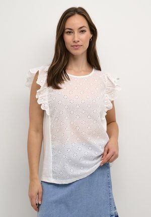 Blusa blanca con hombros con volantes, con patrones de bordado de ojal. Textura suave, escote redondeado y corte fluido. Combinada con falda de mezclilla.