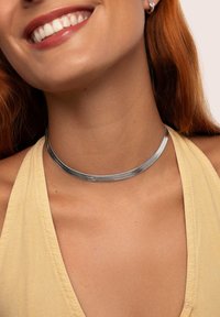 Choker de metal plateado con un diseño liso y plano. Se lleva en el cuello desnudo, combinado con una blusa de color amarillo claro. Apariencia simple y elegante.