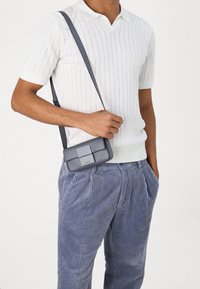 Šedá kabelka přes rameno s hladkou texturou, minimálním čtvercovým klopou a nastavitelným popruhem, nošená s bílým žebrovaným polokošilovým tričkem a světle modrými kordovými kalhotami.