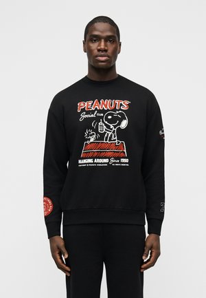 Uomo con felpa nera con grafica a fumetti Peanuts rossa e bianca di Snoopy e Woodstock su una cuccia, che tiene in mano una bevanda.