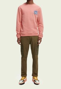 En person som bär en rosa sweatshirt med en liten logotyp, olivgröna cargo-byxor med rutiga lappar, och flerfärgade sneakers med gula snören.