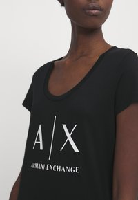 Čierne tričko s krátkymi rukávmi, s hlbokým výstrihom a bielym logom "A/X ARMANI EXCHANGE" viditeľne umiestneným. Mäkká textúra látky.
