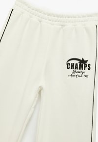 Białe spodnie dresowe z miękkiego materiału, z czarnym nadrukiem logo „CHAMPS Brooklyn Athl. Club 1982” oraz czarnymi paskami po bokach. Elastyczny pas.