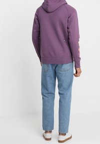 Fialová mikina s kapucí, žebrovaným lemem a logem na rukávu. Kombinováno s světle modrými džíny a bílými teniskami.