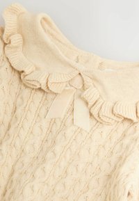 Cream cable-knit sweater with ruffled collar and velvet bow detail at the neckline on a light background.