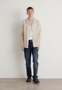 Light beige short-sleeve shirt over a white tank, paired with dark blue jeans and black shoes. The shirt features a button-up design.