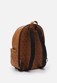 Ruskea reppu mustilla säädettävillä strapseilla ja toistuvalla monogrammikuviolla, näkyy takaa valkoista taustaa vasten.
