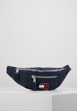 Granatowa nerka z dwoma srebrnymi suwakami i naszywką z logo Tommy Jeans na przedniej kieszeni, prezentowana na białym piedestale.