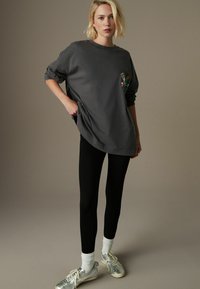 Graues, übergroßes Sweatshirt mit floralem Stickmuster auf der linken Brust, kombiniert mit schwarzen Leggings und silbernen Sportschuhen mit weißen Socken.