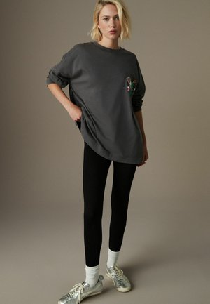 Grijze oversized sweatshirt met bloemenborduursel op de linkerkant van de borst, gecombineerd met zwarte leggings en zilveren sportschoenen met witte sokken.