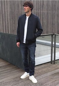 Nuorodotas vyras, vilkintis juodą bomber tipo striukę, baltą marškinėlį, tamsias džinsines kelnes ir baltus sportbačius, stovi ant medinio denio su šiuolaikiniu turėklu fone.