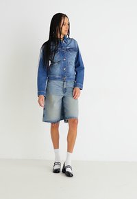 Dżinsowa kurtka z niebieskim wykończeniem, z przednimi kieszeniami i metalowymi guzikami, połączona z dżinsowymi szortami. Model nosi białe skarpetki i czarne buty.