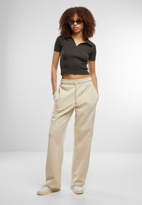 Svart kortermet polo-skjorte, beige joggebukser med elastisk midje, lyse sneakers, modell med krøllete hår og solbriller, står med hendene i lommene.