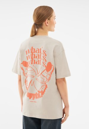 Beige cotton t-shirt featuring orange graphic text and a butterfly design on the back. Short sleeves and a relaxed fit.