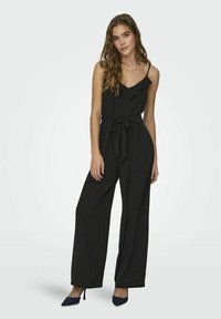 Zwarte jumpsuit met een gerimpelde halslijn, een taillering met riem en een ontwerp met wijde benen. De stof lijkt lichtgewicht en textuur te hebben, gecombineerd met marineblauwe hakschoenen.