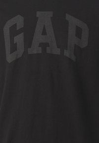 T-shirt noir avec un grand texte graphique en lettres épaisses "GAP" d'une teinte légèrement plus claire sur le devant. Tissu en coton, coupe décontractée.