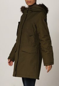Veste parka vert olive avec capuche doublée de fourrure, fermeture éclair frontale et plusieurs poches plaquées pour le rangement. Texture lisse et manches longues.