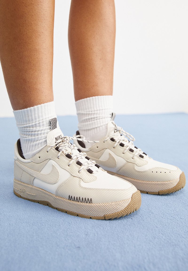 Baskets Nike en mesh beige et blanc avec des accents texturés, œillets noirs et un système de laçage tissé. Semelle extérieure en caoutchouc avec adhérence à motif.