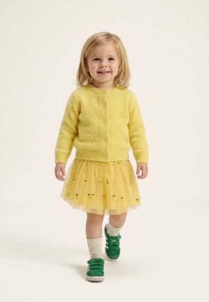 Young child walking forward wearing a yellow cardigan, yellow skirt with lemon patterns, cream socks, and green sneakers.