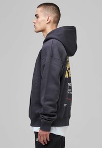 Young man in profile wearing dark oversized hoodie with printed text on back, white shirt underneath, and black pants against gray background.