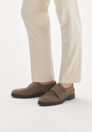 Brązowe zamszowe buty typu slip-on z zaokrąglonym noskiem, z podwójnym paskiem na klamry i przeszyciami, w zestawie z kremowymi spodniami.
