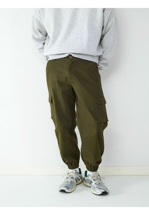 Osoba nosząca oliwkowo-zielone spodnie typu cargo z elastycznymi ściągaczami i szare trampki, stojąca przy białej ścianie.