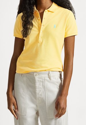 Person iført en gul polo med korte ærmer og fire knapper samt hvide bukser med høj talje, stående foran en ensfarvet baggrund.