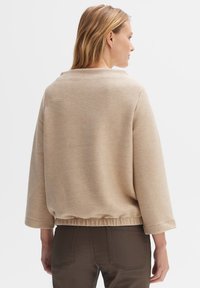 Beige ribbed pullover with three-quarter sleeves, textured fabric, and gathered hem; paired with dark brown pants. Subtle, minimalist design.