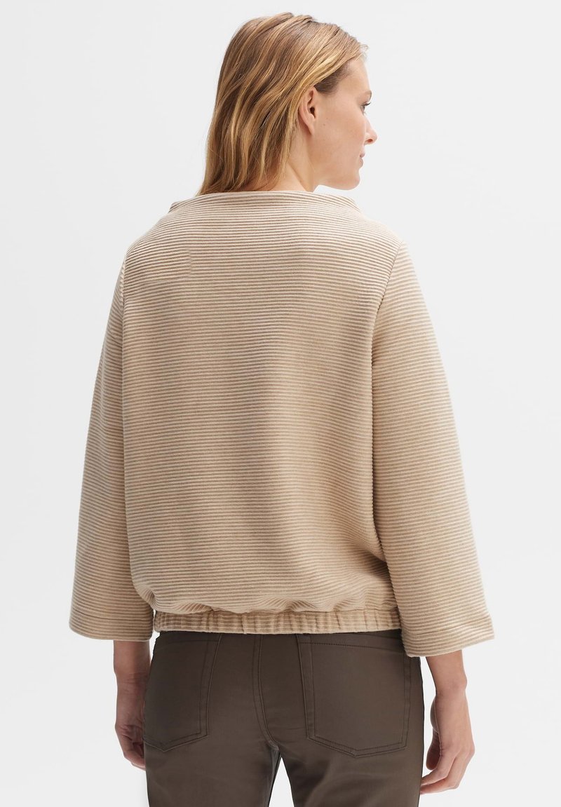Beiger gestreifter Pullover mit Dreiviertelärmeln, strukturiertem Stoff und gerafftem Saum; kombiniert mit dunkelbraunen Hosen. Subtiles, minimalistisches Design.