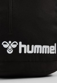Borsa sportiva nera con superficie texture, caratterizzata da un logo bianco con un motivo a forma di ape e la parola "hummel" stampata in modo prominente.