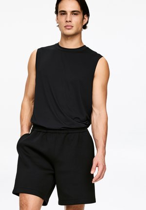 Mann trägt ärmelloses schwarzes Hemd und schwarze Shorts, die Hände in den Taschen vor einem schlichten weißen Hintergrund.