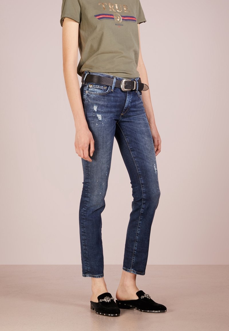 Mörkblå, slitna skinny jeans med hål; kombinerat med en grå T-shirt med guldtext. Svarta slip-on skor med dekorationer.