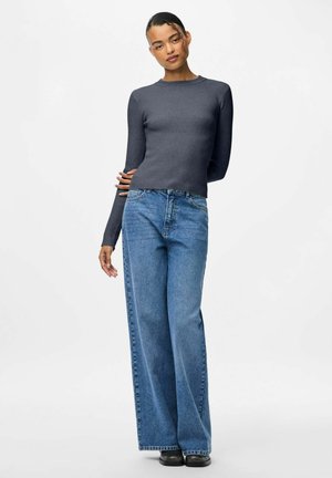 Femme debout avec un bras dans le dos, portant un top ajusté à manches longues gris foncé et un jean bleu à jambes larges avec des chaussures noires.