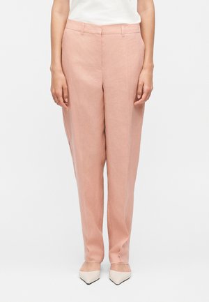 Kvinde iført lyserøde bukser med høj talje og lige ben, kombineret med hvide spidse hæle, stående mod en ensfarvet baggrund.