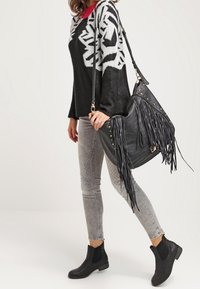 Nainen, jolla on päällään mustavalkoista kuviollista neuletta, harmaat tikkikankaiset farkut, mustat nilkkasaappaat ja hän kantaa suurta mustaa nahkaista fringe-olkalaukkua.