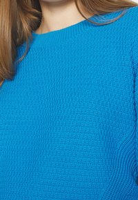 Gros plan sur une personne portant un pull en laine bleu vif, montrant la texture détaillée et une partie de cheveux brun clair.