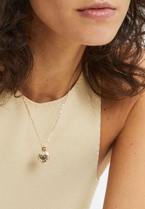 Gold necklace with a geometric heart-shaped pendant, worn on a light beige top. Chain features small links, adding texture and elegance.