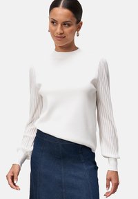 Pull blanc avec des manches longues côtelées et un corps ajusté ; associé à une jupe en denim foncé. La texture est lisse avec un léger éclat.
