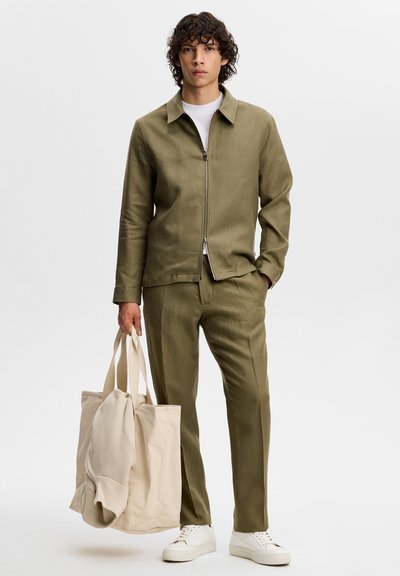 Młody mężczyzna ubrany w oliwkowozieloną kurtkę na suwak i pasujące spodnie, białe trampki, trzyma beżową torbę typu tote z beżowym swetrem w środku.