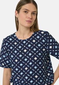 Das marineblaue kurzärmlige Oberteil zeichnet sich durch ein geometrisches Muster mit hellblauen und weißen Formen aus. Der Stoff wirkt weich und hat einen lockeren Sitz.