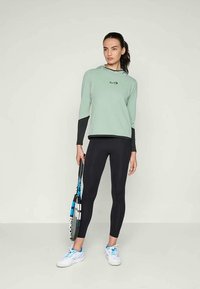 Donna in felpa verde menta e leggings neri, che tiene in mano una racchetta da tennis, in piedi contro uno sfondo chiaro e semplice.