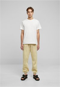 Hvit bomull t-skjorte, gule fleece joggebukser, svarte sandaler med stropper. Modell som står, med hendene langs sidene, enkel grå bakgrunn.