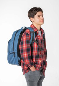 Modrý batoh s texturovanou tkaninou, několika kapsami a polstrovanými ramenními popruhy, který nosí osoba v košili s červeným a černým kostkovaným vzorem.