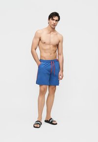 Skjorteløs mann iført blå badebukser med rød snor og svarte slidesandaler, stående mot en ensfarget grå bakgrunn.