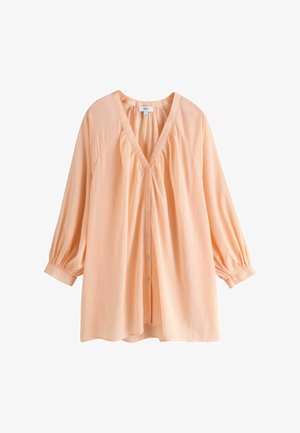 Femme portant une robe chemise ample de couleur pêche claire boutonnée et un collier de perles blanches, debout en extérieur près de la mer, les bras légèrement étendus.