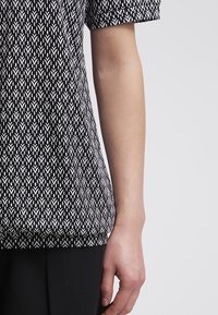 Kortärmad topp i svart och vitt med ett geometriskt mönster. Lättviktsmaterial, lös passform och rundad nederkant.
