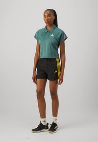 Kobieta stoi w zielonym, krótkim polo, czarnych szortach z żółtymi paskami, białych skarpetkach i czarnych butach sportowych.