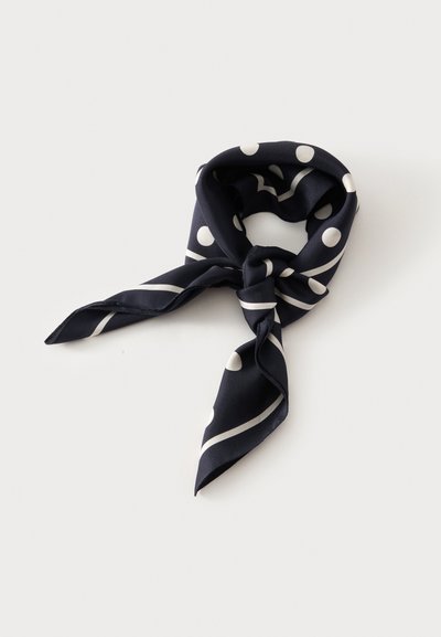 Foulard en soie noire avec des pois blancs et des rayures diagonales, noué lâchement sur fond blanc.