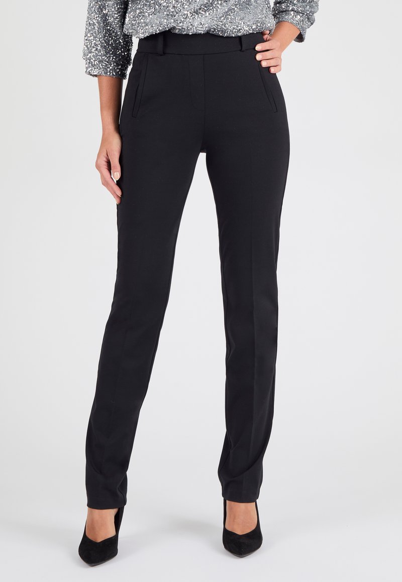 Zwarte, op maat gemaakte broek met een gladde textuur, voorzien van zijzakken en een slim fit. Gecombineerd met zwarte puntige hakken en een glinsterende top.